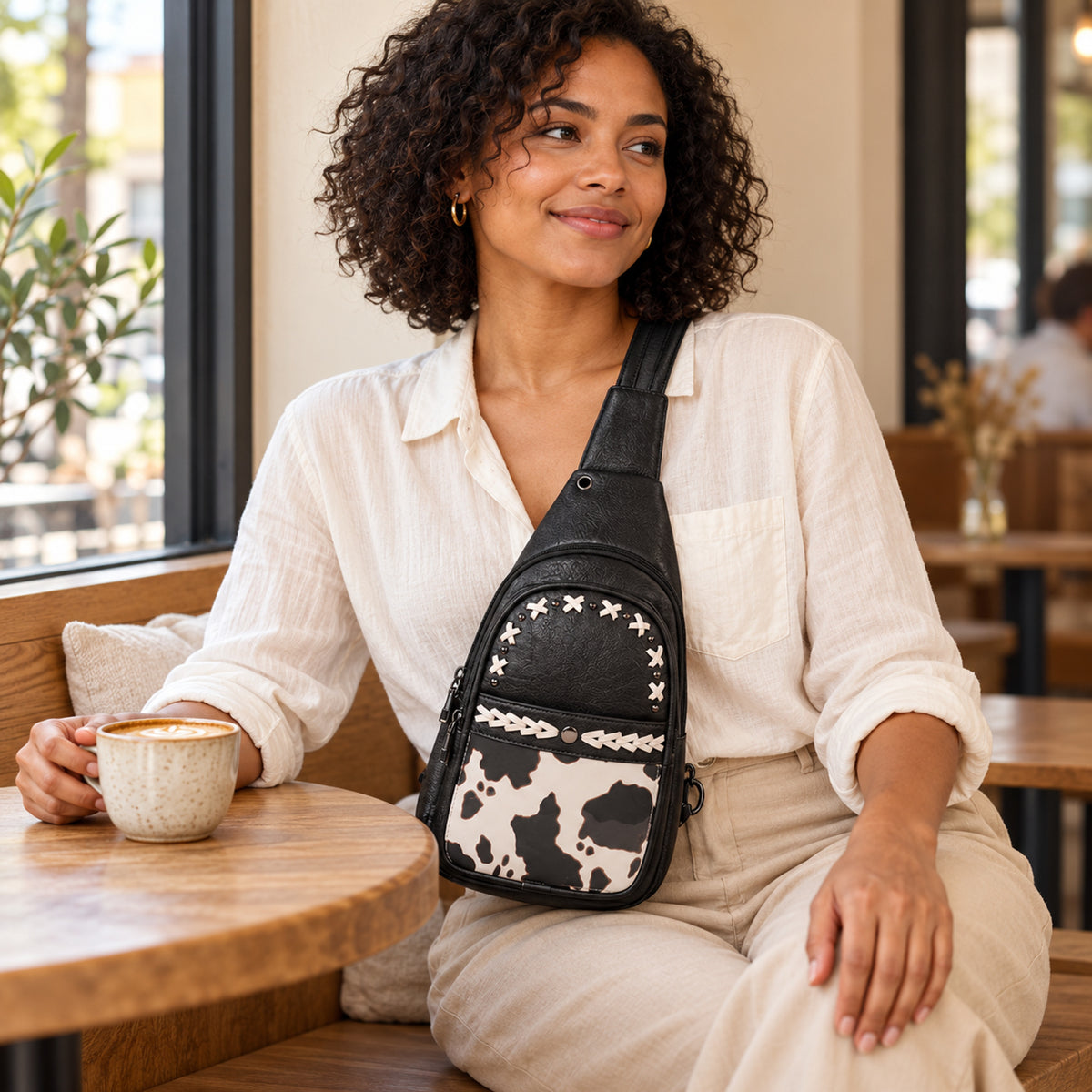 Woman seated at a cafe wearing the Black cow-print convertible bag across the front of her body.